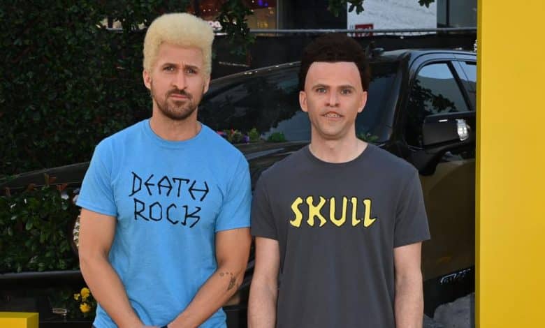 Two men stand in front of a blurred car and floral background. the man on the left has a blond buzz cut and wears a blue shirt labeled "party rock," and the man on the right, sporting short dark hair, wears a black shirt with "skull" printed on it.