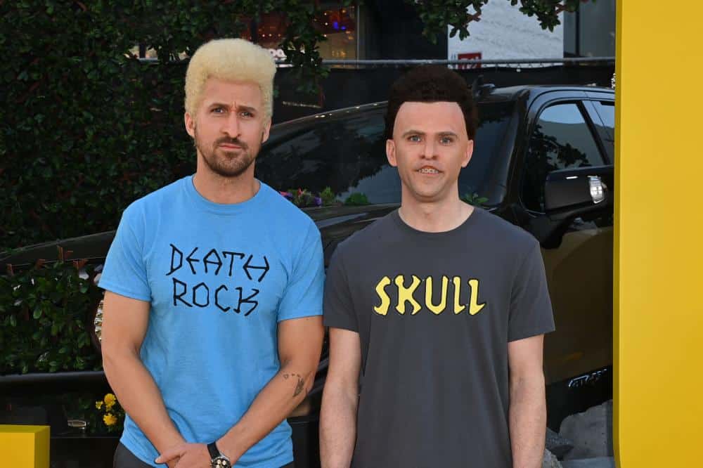 Two men stand in front of a blurred car and floral background. the man on the left has a blond buzz cut and wears a blue shirt labeled "party rock," and the man on the right, sporting short dark hair, wears a black shirt with "skull" printed on it.