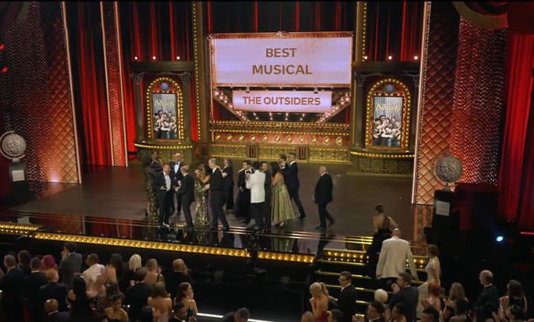 A group of people stands on stage at an awards ceremony under a large screen displaying "Best Musical" and "The Outsiders." The stage is decorated with ornate elements and red curtains. The audience is seated and applauding.