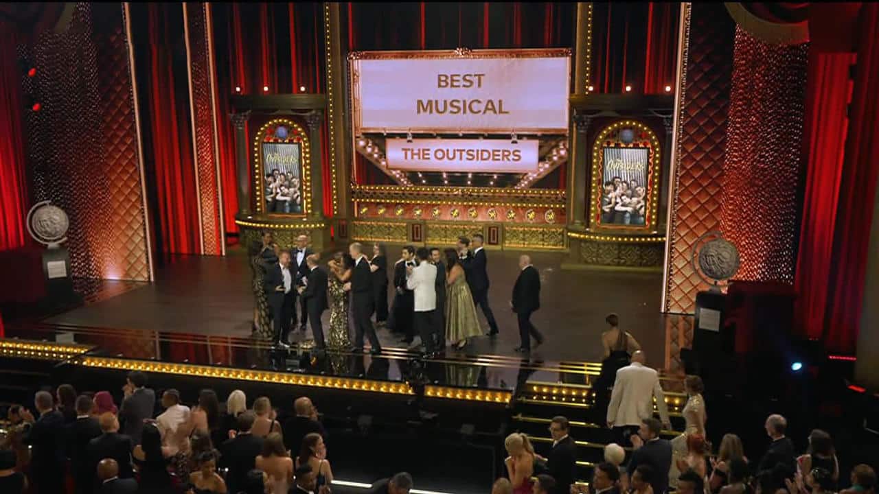 A group of people stands on stage at an awards ceremony under a large screen displaying "Best Musical" and "The Outsiders." The stage is decorated with ornate elements and red curtains. The audience is seated and applauding.