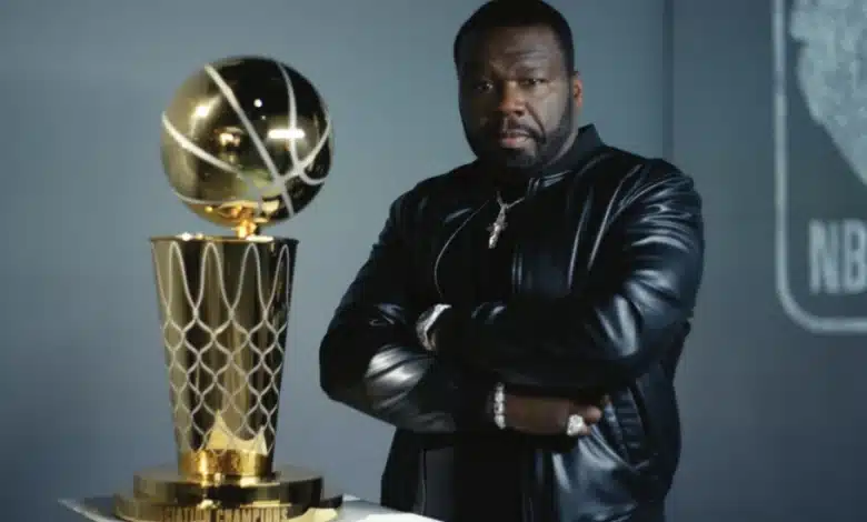 50 Cent, A man with a beard stands with his arms crossed, wearing a black leather jacket and multiple rings. He is next to a large gold trophy featuring a basketball on top, which sits on a pedestal. The background includes an out-of-focus NBA logo.