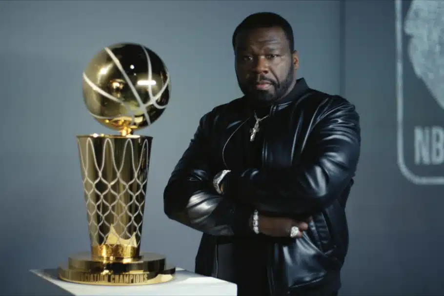 50 Cent, A man with a beard stands with his arms crossed, wearing a black leather jacket and multiple rings. He is next to a large gold trophy featuring a basketball on top, which sits on a pedestal. The background includes an out-of-focus NBA logo.