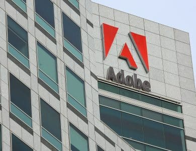 A modern high-rise building features the Adobe logo prominently near its top. The façade consists of gray stone with large, green-tinted glass windows. The Adobe logo shows a red stylized "A" above the word "Adobe" written in gray. The sky is clear, presenting a clean, corporate aesthetic.