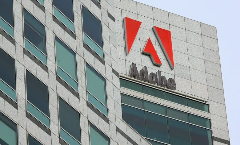 A modern high-rise building features the Adobe logo prominently near its top. The façade consists of gray stone with large, green-tinted glass windows. The Adobe logo shows a red stylized "A" above the word "Adobe" written in gray. The sky is clear, presenting a clean, corporate aesthetic.