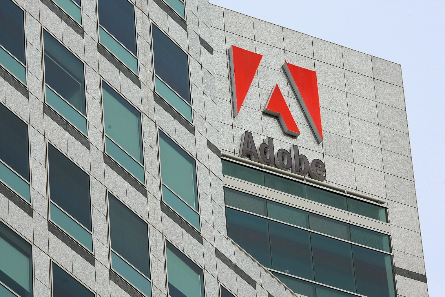 A modern high-rise building features the Adobe logo prominently near its top. The façade consists of gray stone with large, green-tinted glass windows. The Adobe logo shows a red stylized "A" above the word "Adobe" written in gray. The sky is clear, presenting a clean, corporate aesthetic.