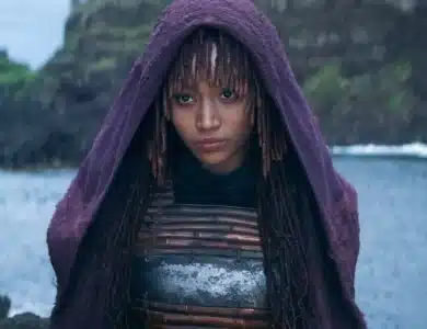 Amandla Stenberg Mae, A person with long, braided hair and bangs stands by a body of water with cliffs in the background. They are wearing a purple hooded cloak and a metallic chest plate. The cloak partially obscures their face, emphasizing their serious expression and contemplative gaze towards the camera.