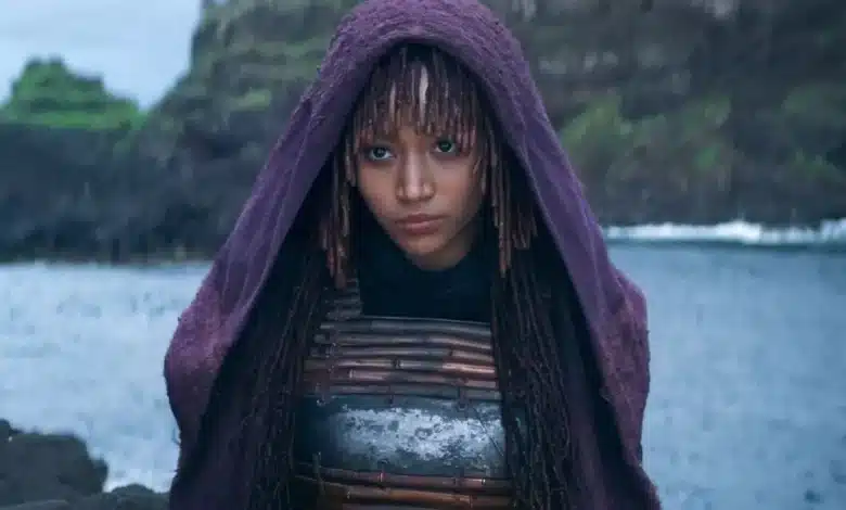 Amandla Stenberg Mae, A person with long, braided hair and bangs stands by a body of water with cliffs in the background. They are wearing a purple hooded cloak and a metallic chest plate. The cloak partially obscures their face, emphasizing their serious expression and contemplative gaze towards the camera.