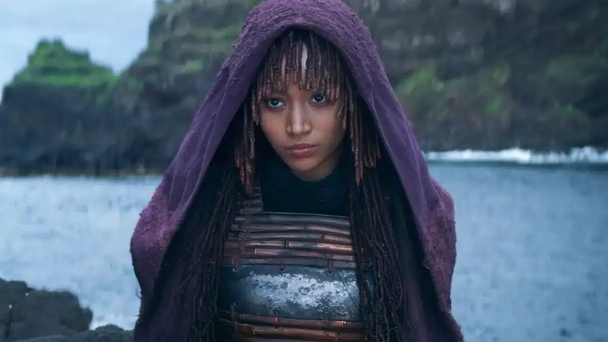 Amandla Stenberg Mae, A person with long, braided hair and bangs stands by a body of water with cliffs in the background. They are wearing a purple hooded cloak and a metallic chest plate. The cloak partially obscures their face, emphasizing their serious expression and contemplative gaze towards the camera.