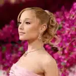 Ariana Grande, A person with light skin, blonde hair styled in a wavy ponytail, wears a strapless pink dress and drop earrings. They are standing against a backdrop of vibrant pink flowers, looking confidently sideways while smiling softly. A diamond necklace complements their elegant appearance.