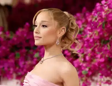 Ariana Grande, A person with light skin, blonde hair styled in a wavy ponytail, wears a strapless pink dress and drop earrings. They are standing against a backdrop of vibrant pink flowers, looking confidently sideways while smiling softly. A diamond necklace complements their elegant appearance.