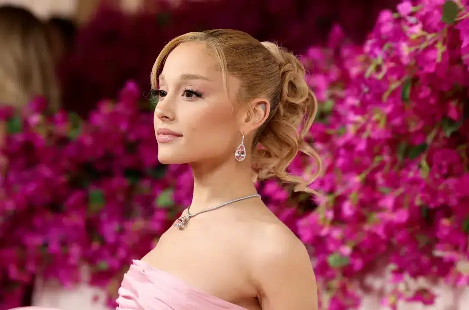 Ariana Grande, A person with light skin, blonde hair styled in a wavy ponytail, wears a strapless pink dress and drop earrings. They are standing against a backdrop of vibrant pink flowers, looking confidently sideways while smiling softly. A diamond necklace complements their elegant appearance.