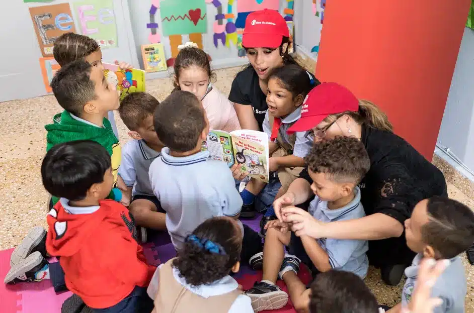 Camilla Cabello & Two women, each wearing a red cap and black shirts, read a book to a group of young children who are gathered around them. The children are sitting on a colorful play mat in a classroom. The background shows educational posters, colorful walls, and various classroom decorations.