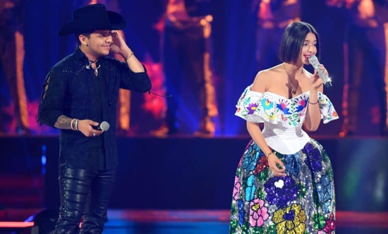 Christian Nodal and Angela Aguilar, A man in a black outfit and a cowboy hat stands beside a woman singing into a microphone. She wears a white off-the-shoulder blouse and a colorful, floral-patterned skirt. Both are on a brightly lit stage, and blurred musicians play instruments in the background.
