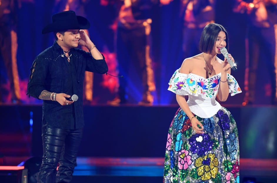Christian Nodal and Angela Aguilar, A man in a black outfit and a cowboy hat stands beside a woman singing into a microphone. She wears a white off-the-shoulder blouse and a colorful, floral-patterned skirt. Both are on a brightly lit stage, and blurred musicians play instruments in the background.