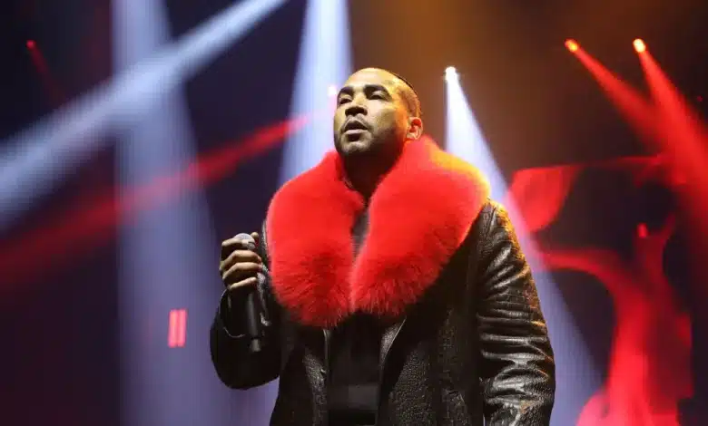 Don Omar, A person is performing on stage, holding a microphone. They are wearing a black leather jacket with a large, bright red fur collar. The background is illuminated with red and white stage lights, creating a dramatic and vibrant atmosphere.