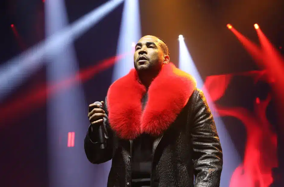 Don Omar, A person is performing on stage, holding a microphone. They are wearing a black leather jacket with a large, bright red fur collar. The background is illuminated with red and white stage lights, creating a dramatic and vibrant atmosphere.