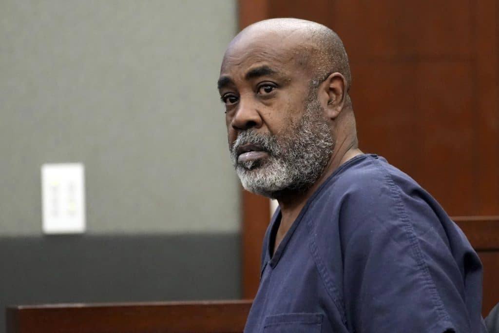 Duane "Keffe-D" Davis, A serious-looking man with a bald head and gray beard, wearing a dark blue prison uniform, sits in a courtroom. He gazes intently towards the camera. The background shows wood paneling and a gray wall with an electrical outlet visible.