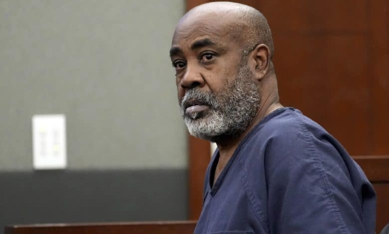 Duane "Keffe-D" Davis, A serious-looking man with a bald head and gray beard, wearing a dark blue prison uniform, sits in a courtroom. He gazes intently towards the camera. The background shows wood paneling and a gray wall with an electrical outlet visible.