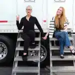 Jamie Lee Curtis and Lindsay Lohan, Two women pose outdoors, sitting on metal steps attached to a white trailer. The woman on the left, with gray hair and glasses, wears a black outfit, making a playful hand gesture with her tongue out. The woman on the right, with long red hair, wears a striped sweater and jeans, smiling warmly.