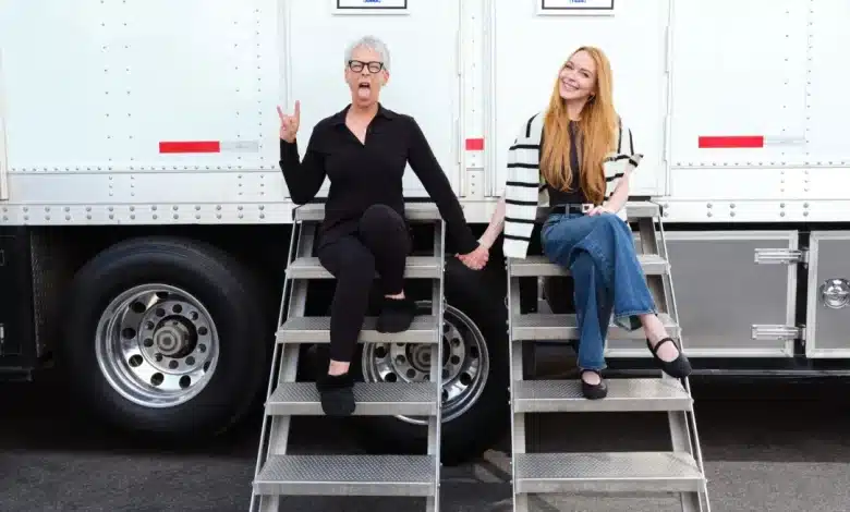 Jamie Lee Curtis and Lindsay Lohan, Two women pose outdoors, sitting on metal steps attached to a white trailer. The woman on the left, with gray hair and glasses, wears a black outfit, making a playful hand gesture with her tongue out. The woman on the right, with long red hair, wears a striped sweater and jeans, smiling warmly.