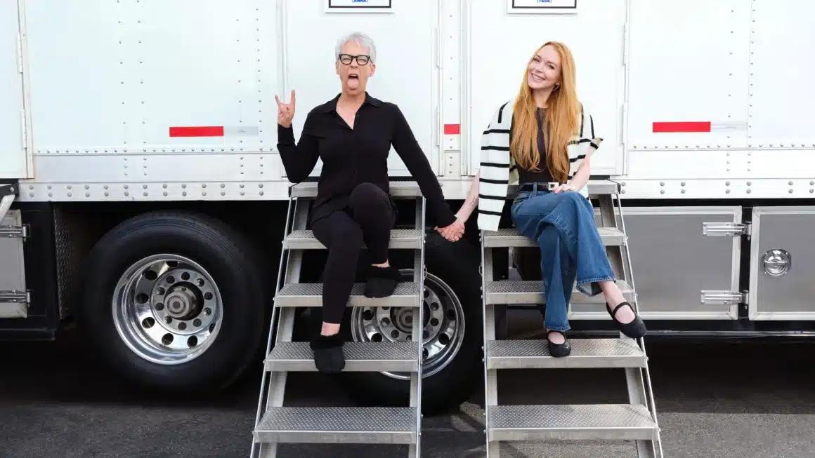 Jamie Lee Curtis and Lindsay Lohan, Two women pose outdoors, sitting on metal steps attached to a white trailer. The woman on the left, with gray hair and glasses, wears a black outfit, making a playful hand gesture with her tongue out. The woman on the right, with long red hair, wears a striped sweater and jeans, smiling warmly.