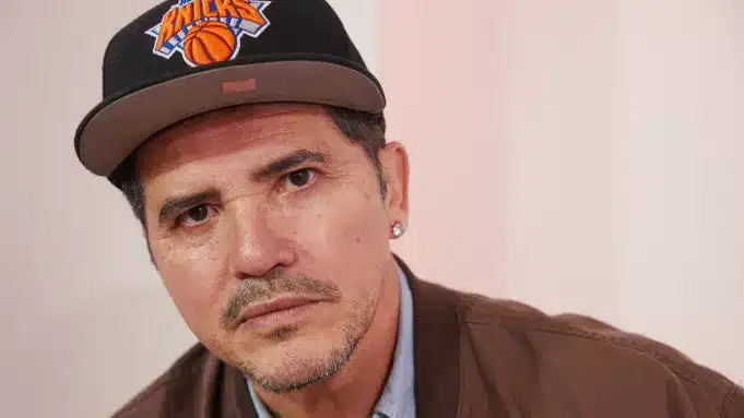 John Leguizamo, A man with a serious expression is wearing a New York Knicks cap, a brown jacket, and a light blue shirt. He has a goatee and an earring in his left ear. The background is a plain light color.