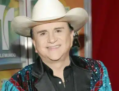 Johnny Canales A person is smiling while wearing a white cowboy hat, a black shirt, and a colorful jacket adorned with sequins. The background features a logo that partially reads "Telemundo" and is composed of various colors including green, yellow, and red.