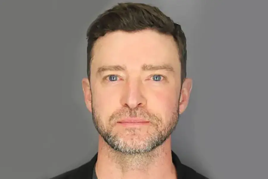 Justin Timberlake, A man with short, dark hair and a trimmed beard is looking directly at the camera with a neutral expression. He is wearing a dark shirt, and the background is plain and gray, focusing attention on his facial features.