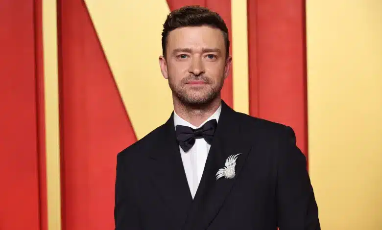 A man with short, brown hair and a trimmed beard is dressed in a formal black suit with a black bow tie and a white bird-shaped brooch. He stands against a red and yellow geometric background, looking directly at the camera.