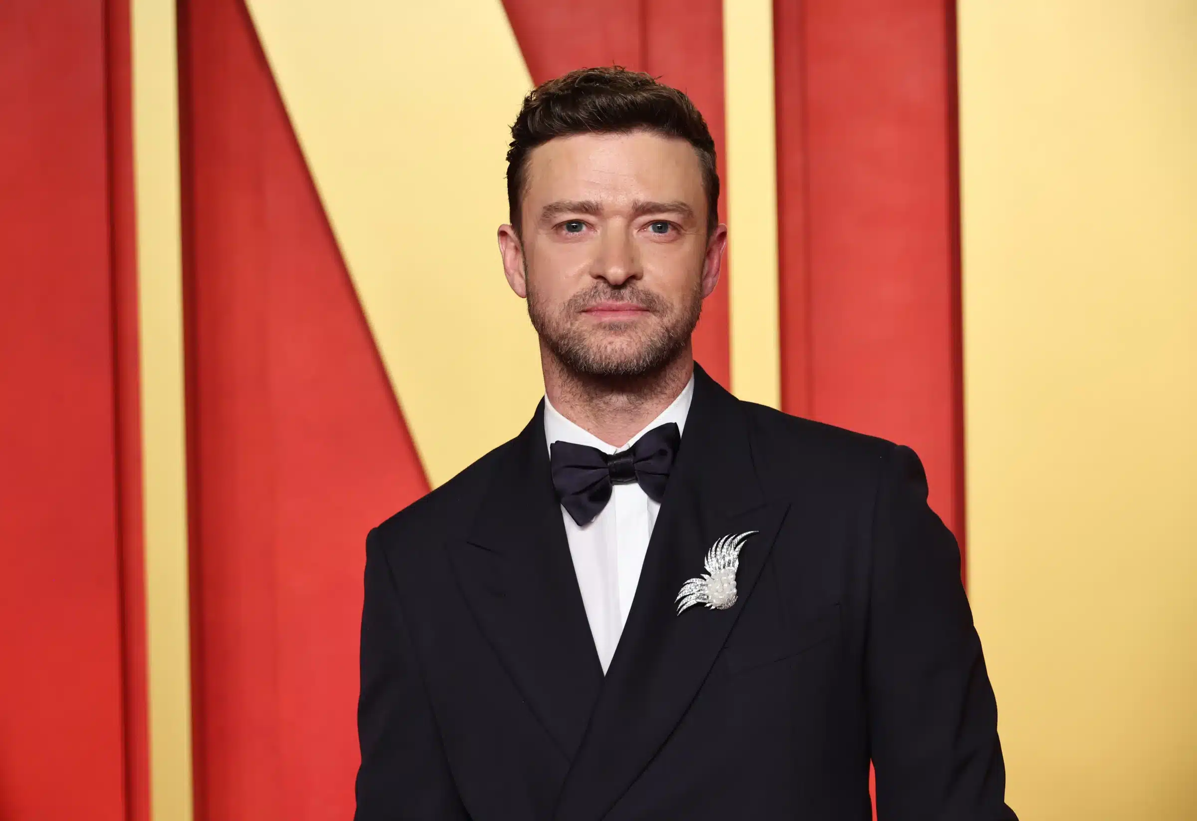A man with short, brown hair and a trimmed beard is dressed in a formal black suit with a black bow tie and a white bird-shaped brooch. He stands against a red and yellow geometric background, looking directly at the camera.
