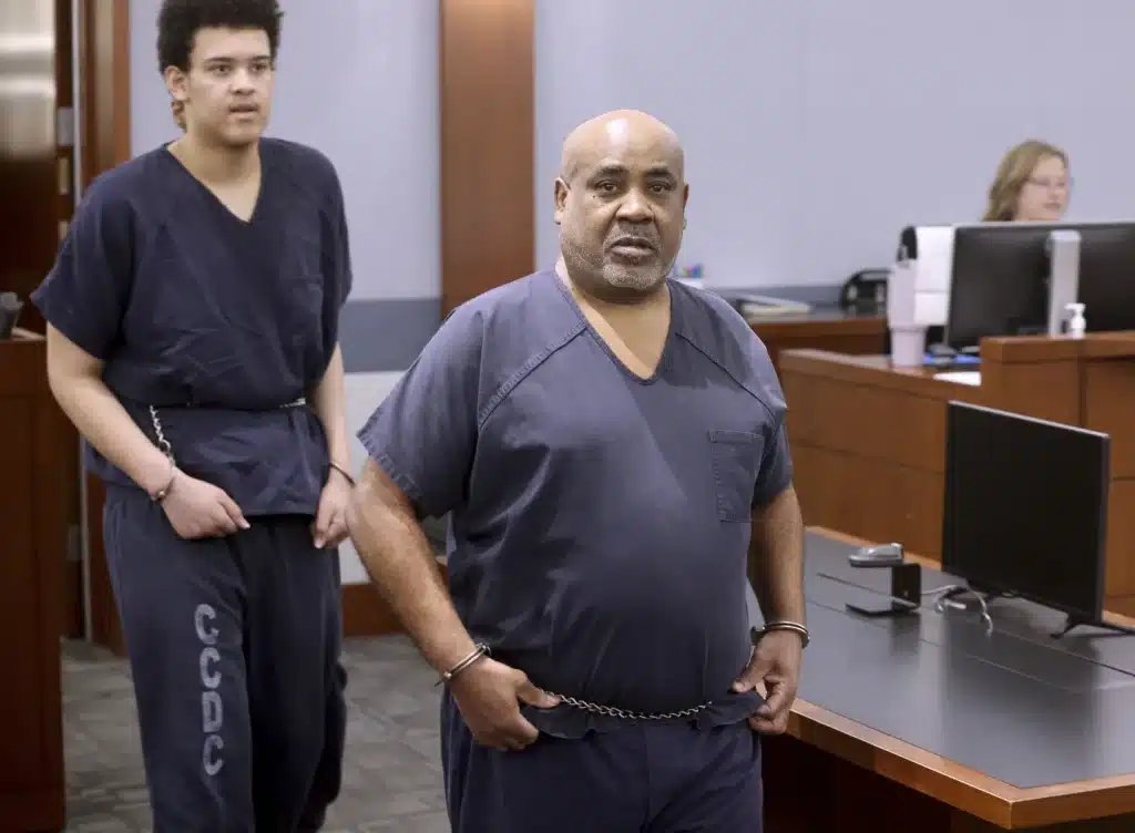 Two men in dark blue prison uniforms and wrist restraints stand in a courtroom. The older man in the foreground has a bald head and a gray beard. The younger man behind him has curly hair. They are accompanied by an officer, and a seated person works on a computer in the background.