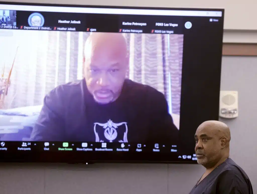 A man in a dark shirt appears on a large monitor during a video conference call. In the foreground, another man wearing scrubs stands facing the screen. The background shows a plain wall and part of a window. The screen displays various icons and participants' names at the bottom.