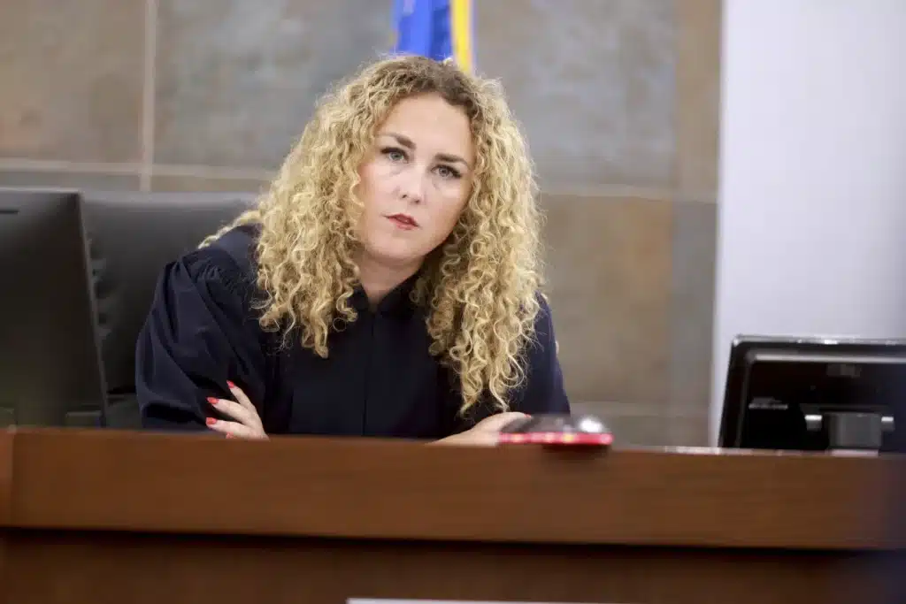 A woman judge with curly blonde hair wearing a black robe sits behind a wooden bench in a courtroom. Her arms are crossed, and she looks directly ahead with a serious expression. A monitor and a flag are visible in the background.