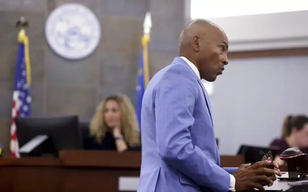 A man in a light blue suit speaks at a podium in a courtroom. Behind him, a woman with long blonde hair sits at a desk with a computer, and another individual is partially visible in the background. The room features flags and a circular emblem on a wall.