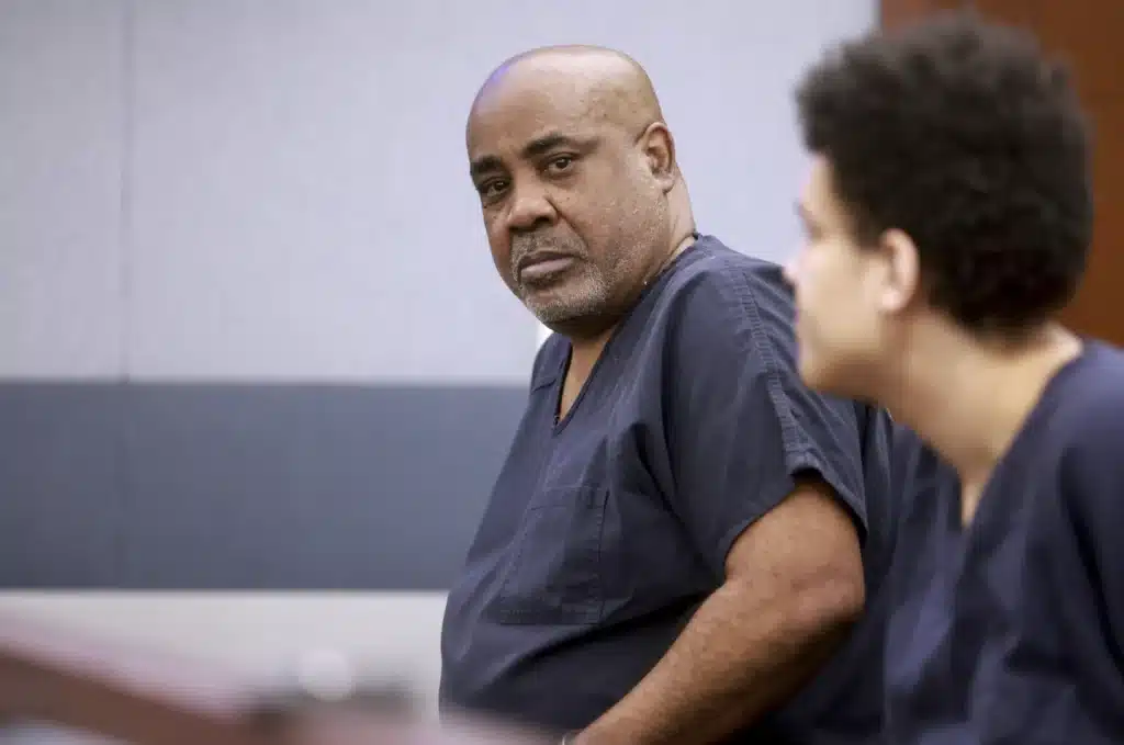 Two men wearing dark-colored prison uniforms are seated in a courtroom. The man in the foreground has a bald head and is looking towards his right. The man in the background has curly hair and is also looking to his right. Both appear to be engaged in the court proceedings.