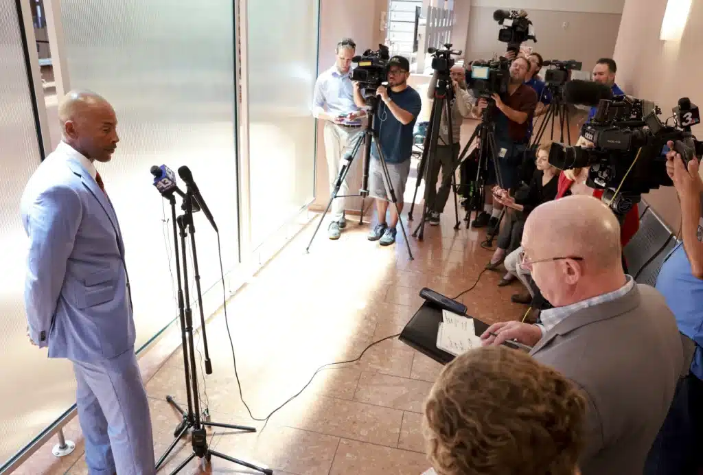 A man in a light blue suit addresses a group of journalists and media personnel in a well-lit indoor setting. Several microphones are positioned in front of him as reporters with cameras, notebooks, and recording devices capture the scene. The audience is attentive, listening intently to his speech.