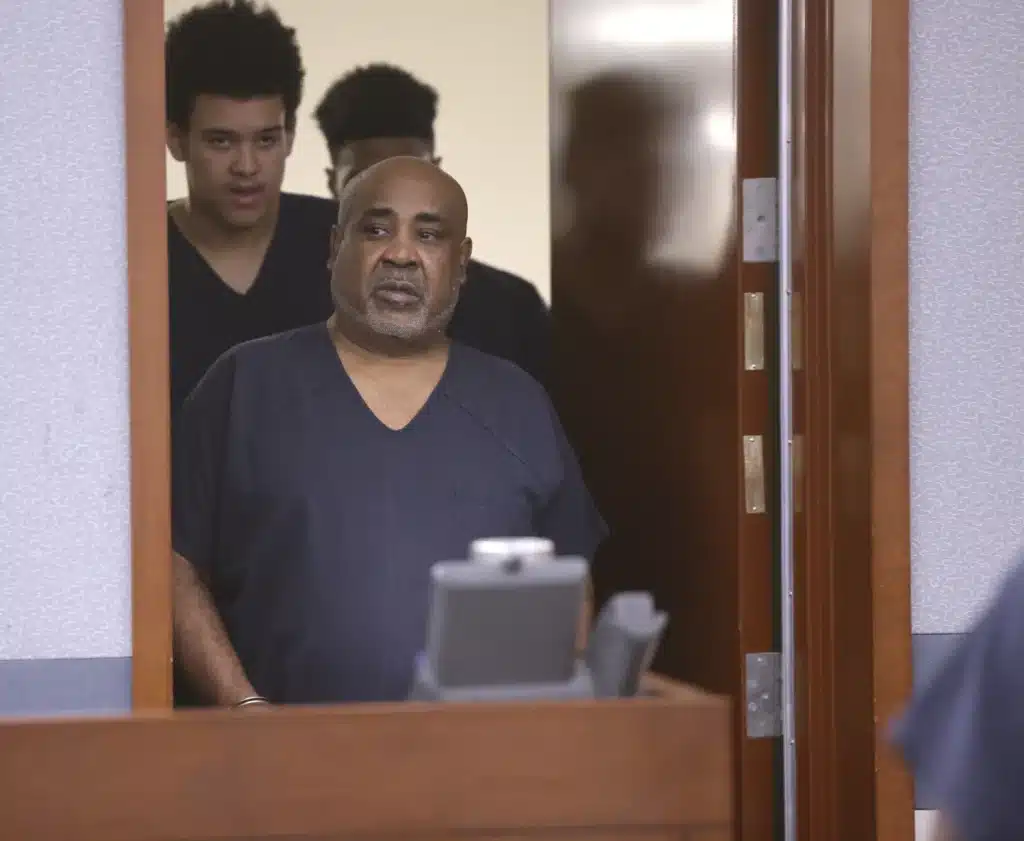 Three men in dark blue prison uniforms with serious expressions stand in a court room. The older bald man with a gray beard is closest to the door, with two younger men behind him. They look towards the front of the room and are framed by the partially open door.