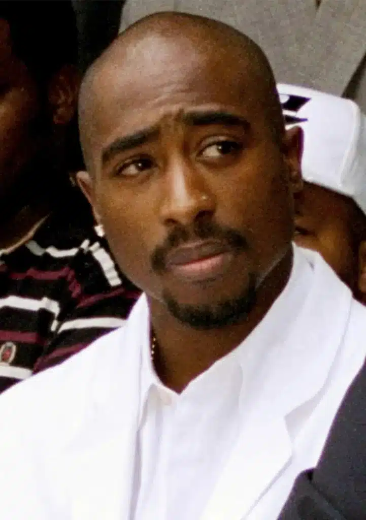 Tupac Shakur, A man with closely shaved hair and a trimmed goatee is wearing a white collared shirt and a white jacket. He appears contemplative and is looking slightly to his left. The background shows other people, including one individual in a striped shirt and another in a white cap.