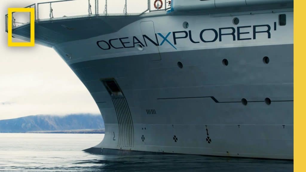 The image shows the starboard bow of a large research vessel named "OceanXplorer," with part of the National Geographic logo visible in the top left corner. The ship is grey and anchored in calm waters with a distant mountainous landscape under a cloudy sky.