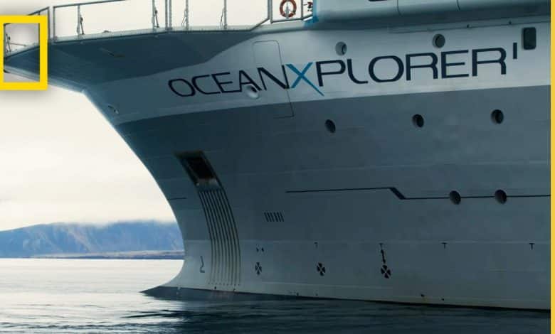 The image shows the starboard bow of a large research vessel named "OceanXplorer," with part of the National Geographic logo visible in the top left corner. The ship is grey and anchored in calm waters with a distant mountainous landscape under a cloudy sky.