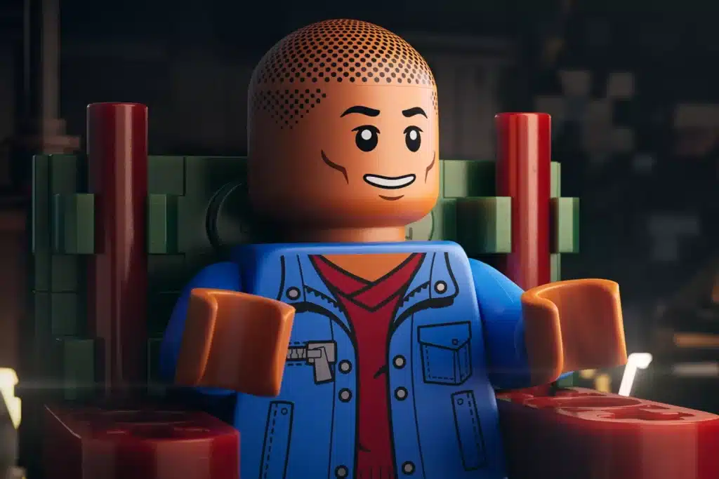 A close-up of a LEGO minifigure sitting on a red and green chair. The minifigure has a smiling face, a bald head with dotted texture, and is wearing a blue jacket over a red shirt. The background is dimly lit, featuring blurred shapes and objects, creating a cozy, atmospheric setting.