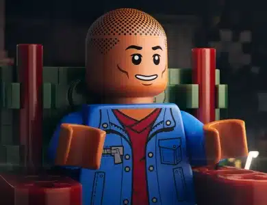 A close-up of a LEGO minifigure sitting on a red and green chair. The minifigure has a smiling face, a bald head with dotted texture, and is wearing a blue jacket over a red shirt. The background is dimly lit, featuring blurred shapes and objects, creating a cozy, atmospheric setting.