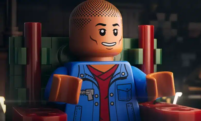 A close-up of a LEGO minifigure sitting on a red and green chair. The minifigure has a smiling face, a bald head with dotted texture, and is wearing a blue jacket over a red shirt. The background is dimly lit, featuring blurred shapes and objects, creating a cozy, atmospheric setting.
