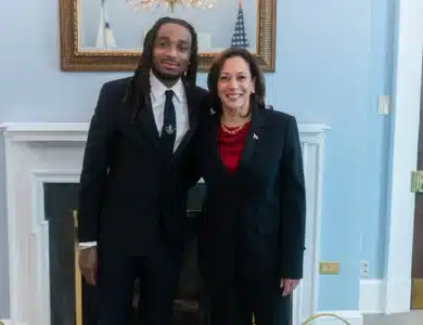Quavo and Vice President Kamala Harris, A man with long dreadlocks wearing a black suit and a woman with shoulder-length hair in a black suit with a red top stand side by side, smiling. They are in front of a white fireplace with framed pictures and a blue wall. The woman has her arm around the man's back.