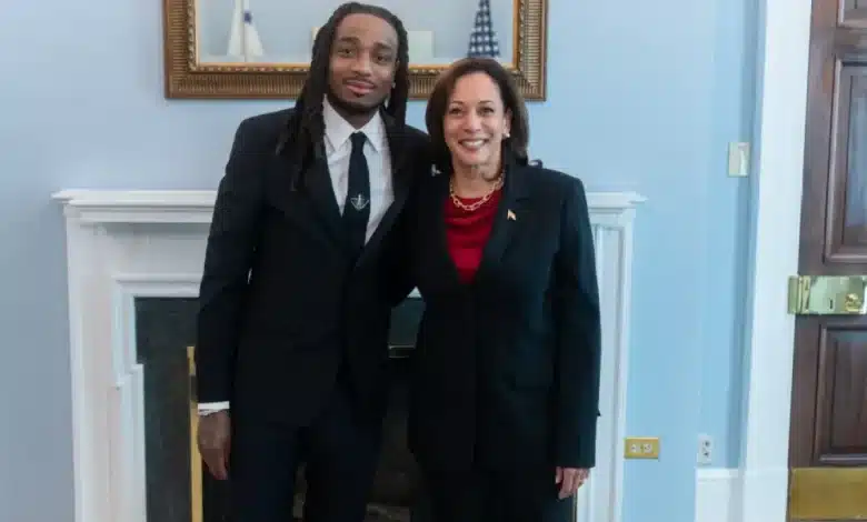 Quavo and Vice President Kamala Harris, A man with long dreadlocks wearing a black suit and a woman with shoulder-length hair in a black suit with a red top stand side by side, smiling. They are in front of a white fireplace with framed pictures and a blue wall. The woman has her arm around the man's back.