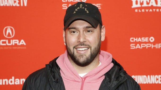 Scooter Braun, A man with a beard and mustache is smiling in front of a red background with white logos and text. He is wearing a black baseball cap with yellow text, a black jacket, and a pink hoodie. The background includes logos for Sundance, Acura, and Chase Sapphire.