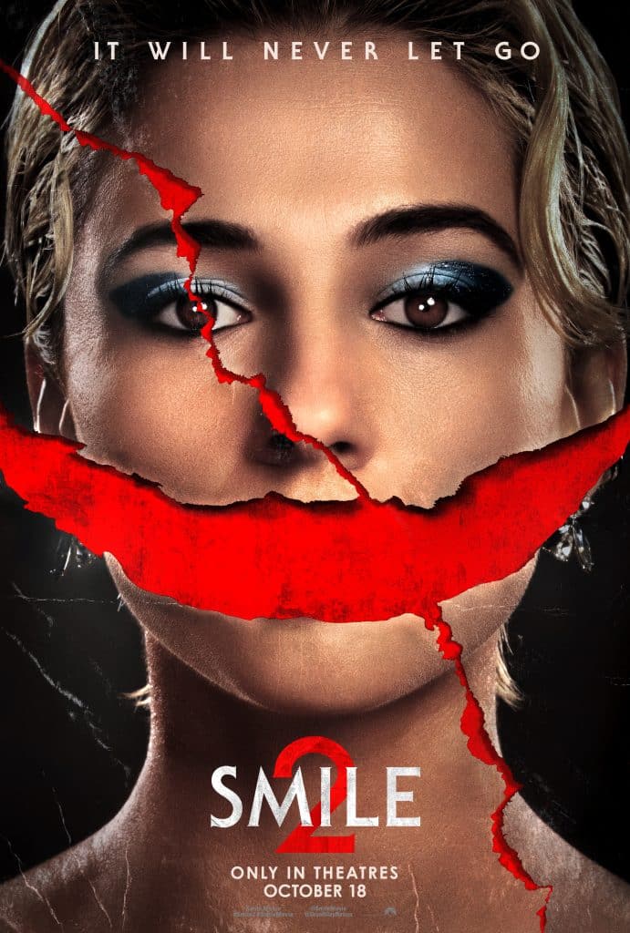 Promotional poster for the movie "Smile 2" featuring a close-up of a woman's face with intense eye makeup. A wide, red, unsettling smile is scratched across her face. The poster text reads “It will never let go” at the top and “Smile 2, Only in Theatres, October 18” at the bottom.