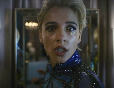 A person with short blonde hair, wearing a sparkly blue and black outfit, is looking directly at the camera with a surprised expression. Teardrop makeup adorns their face, and they are standing in front of a mirror with two lit lamps on either side.