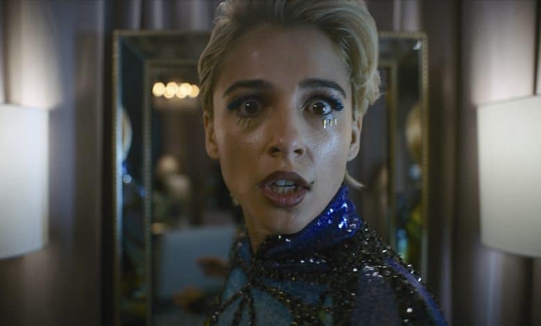 A person with short blonde hair, wearing a sparkly blue and black outfit, is looking directly at the camera with a surprised expression. Teardrop makeup adorns their face, and they are standing in front of a mirror with two lit lamps on either side.