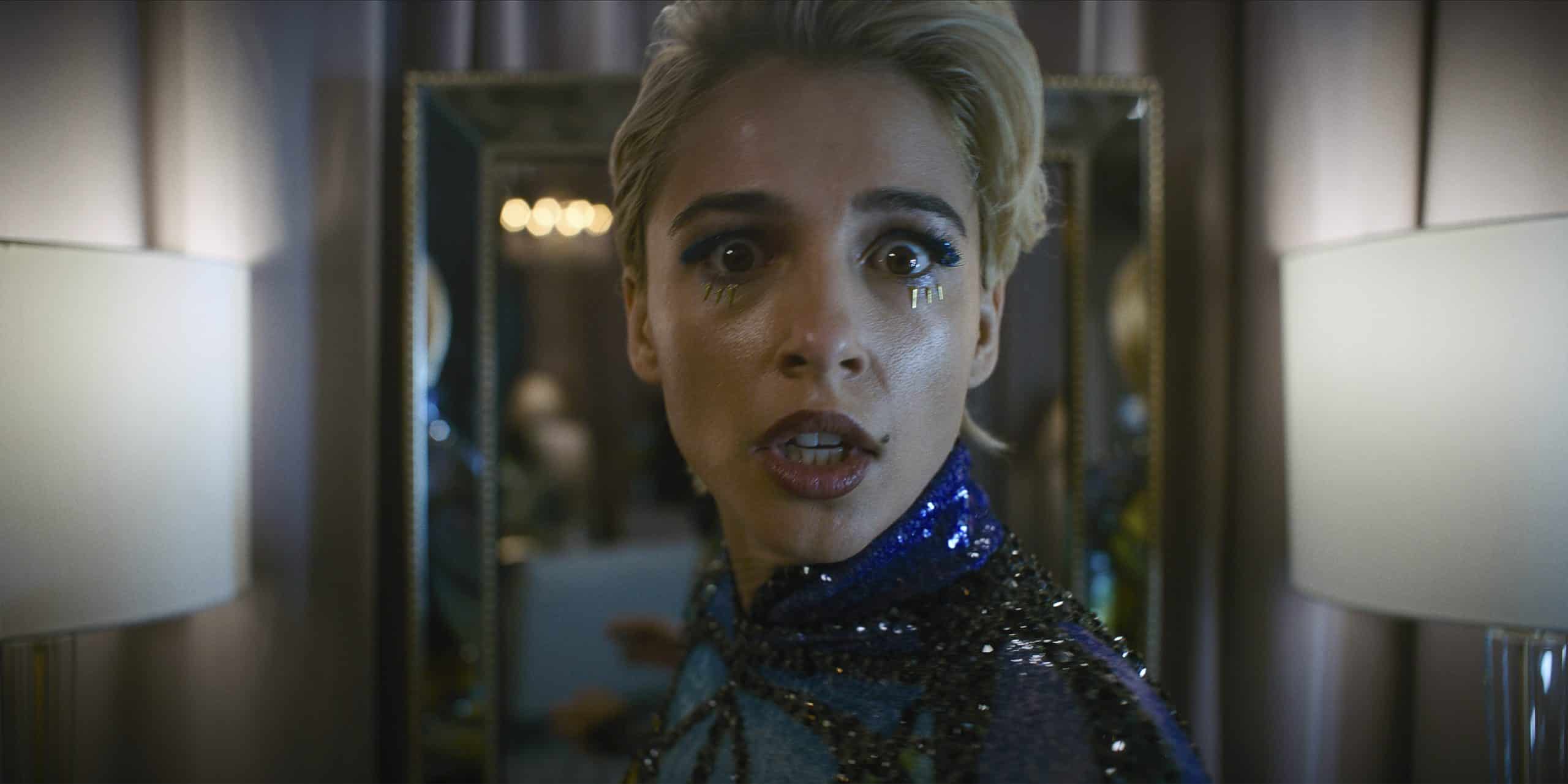 A person with short blonde hair, wearing a sparkly blue and black outfit, is looking directly at the camera with a surprised expression. Teardrop makeup adorns their face, and they are standing in front of a mirror with two lit lamps on either side.